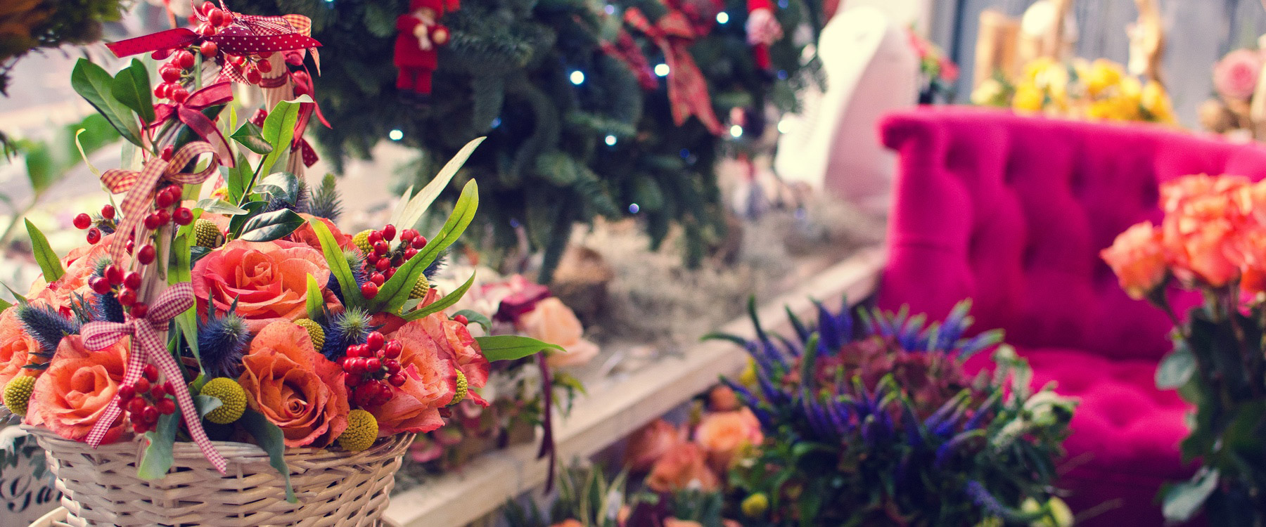 A white wicker basket with an arrangement of peach roses, red berries, and greenery placed indoors near a decorated Christmas tree, surrounded by flowers and a bright pink tufted armchair.