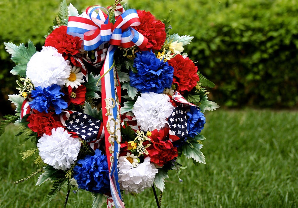 A wreath composed of red, white, and blue flowers with a striped bow and ribbon, set against green grass and a trimmed hedge backdrop.