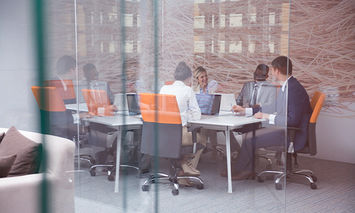 Business professionals having a meeting at a rectangular table with laptops, seen through a glass wall. The modern office has orange-accented chairs and a large abstract mural on the back wall.